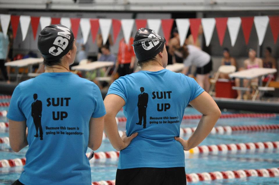 two swimmers with caps and team shirts on facing a competition pool